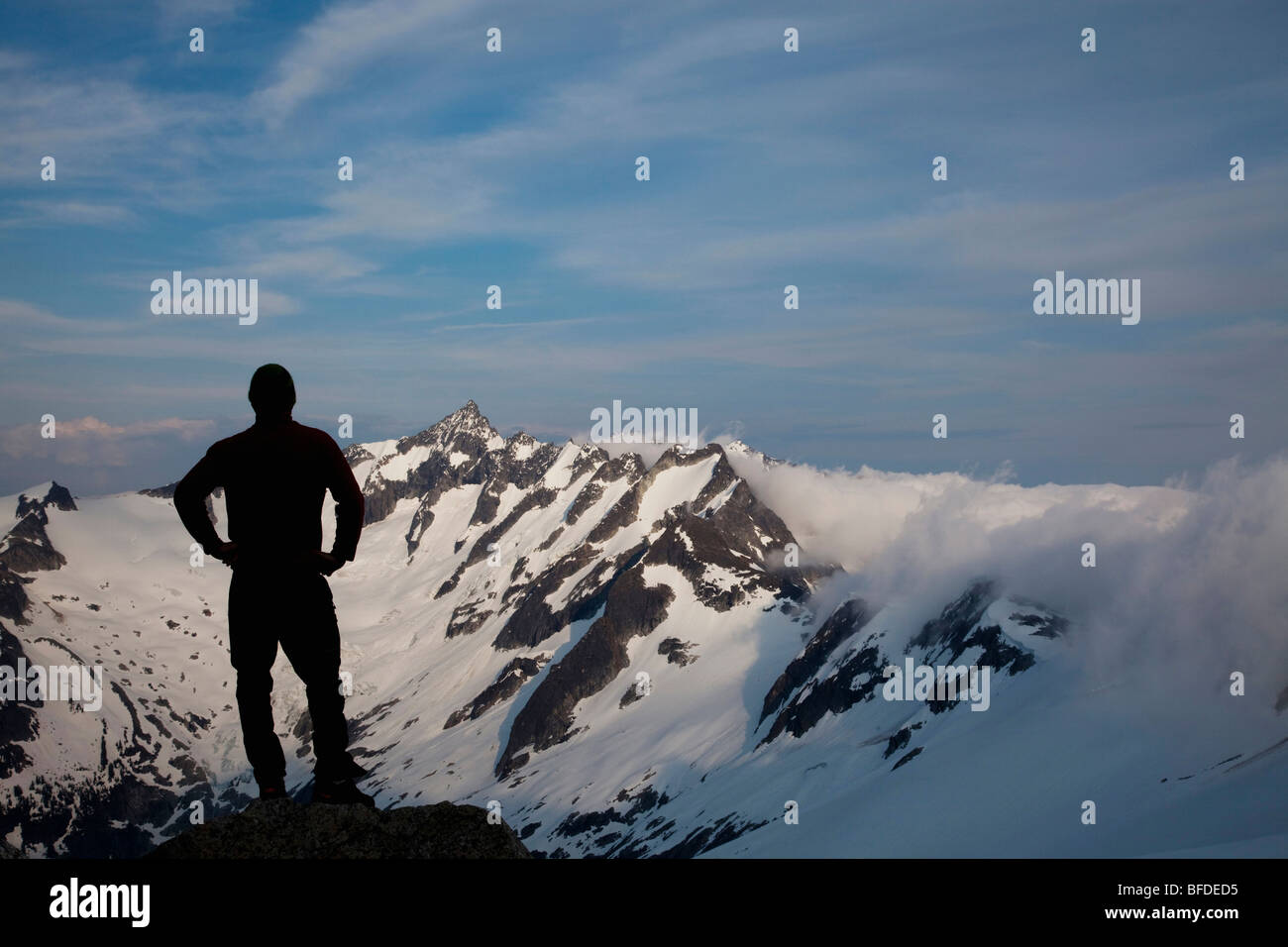 Silhouetted against dramatic backdrop, a young climber stands with arms on his hips silhouetted against a mountain background. Stock Photo