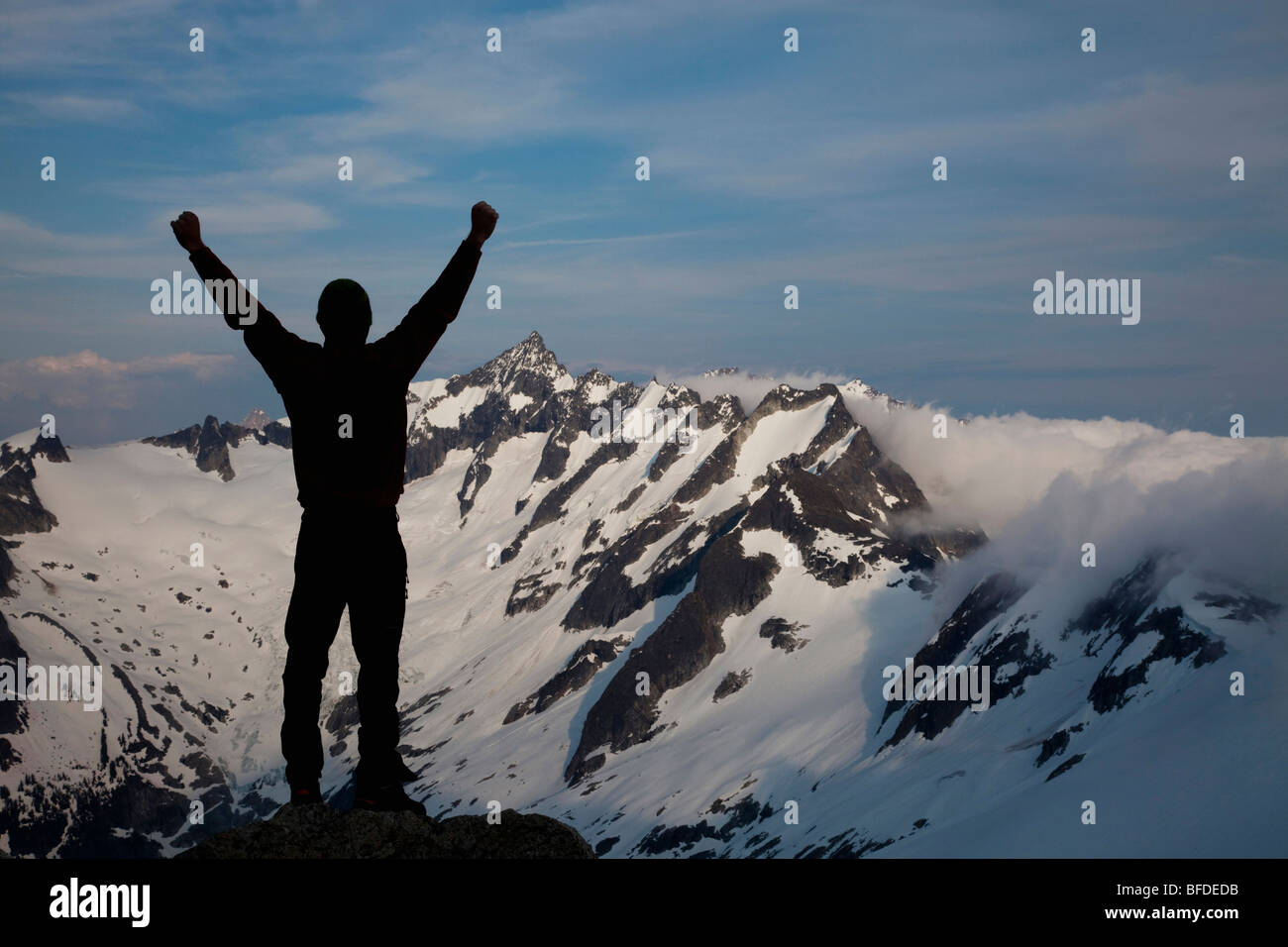 Silhouetted against a dramatic backdrop, a young climber raises his arms while enjoying the mountain view. Stock Photo