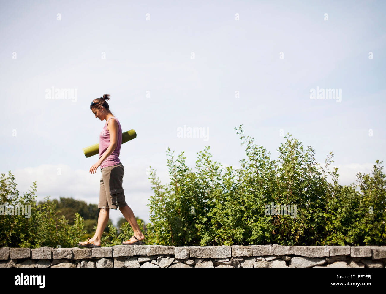 A young woman walks on a stone ledge holding a green yoga mat. Stock Photo