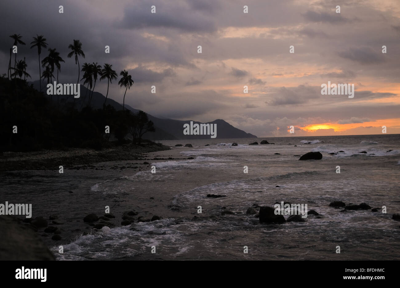 The sun sets over a rocky beach lined with mountains and palm trees in Puerto Colombia, Venezuela. Stock Photo