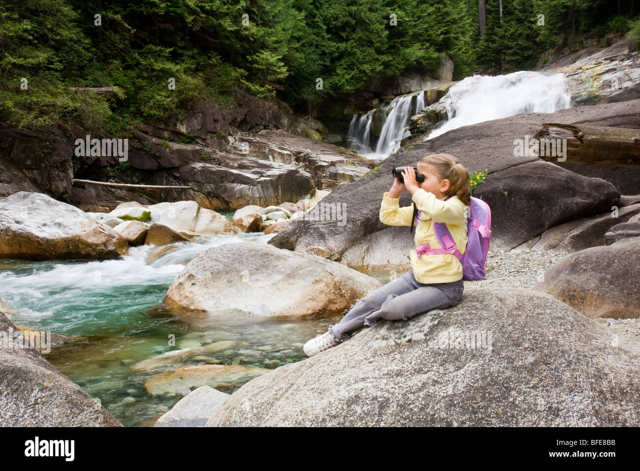 Young girl sitting on a rock and looking through binoculars in front of waterfalls in Golden Ears Provincial Park in Maple Ridge Stock Photo