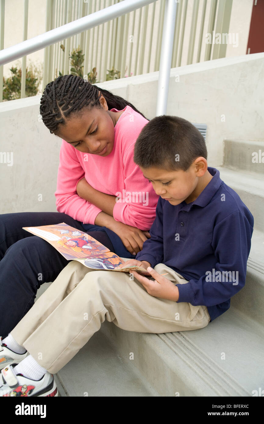 child helping another child Multi ethnic ethnically diverse African American teen girl reads a book to an Hispanic boy on steps. California MR © Myrleen Pearson Stock Photo