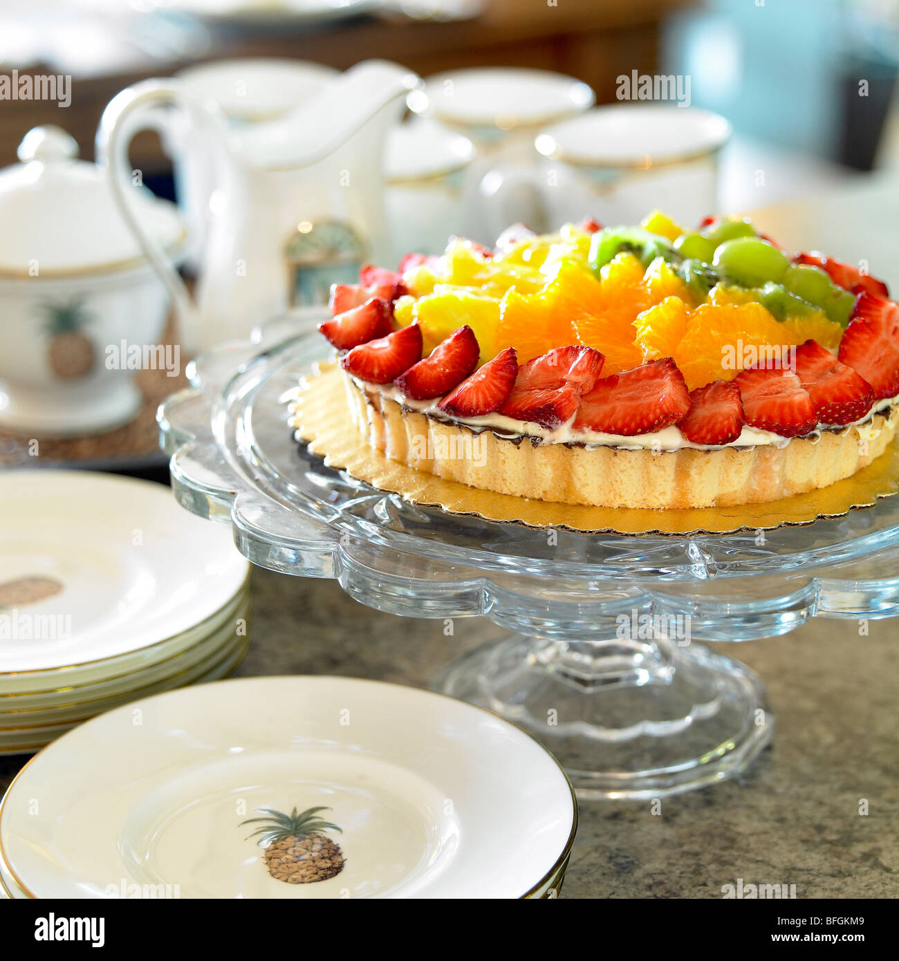 Fruit flan on glass pedestal with tea set in the background Stock Photo