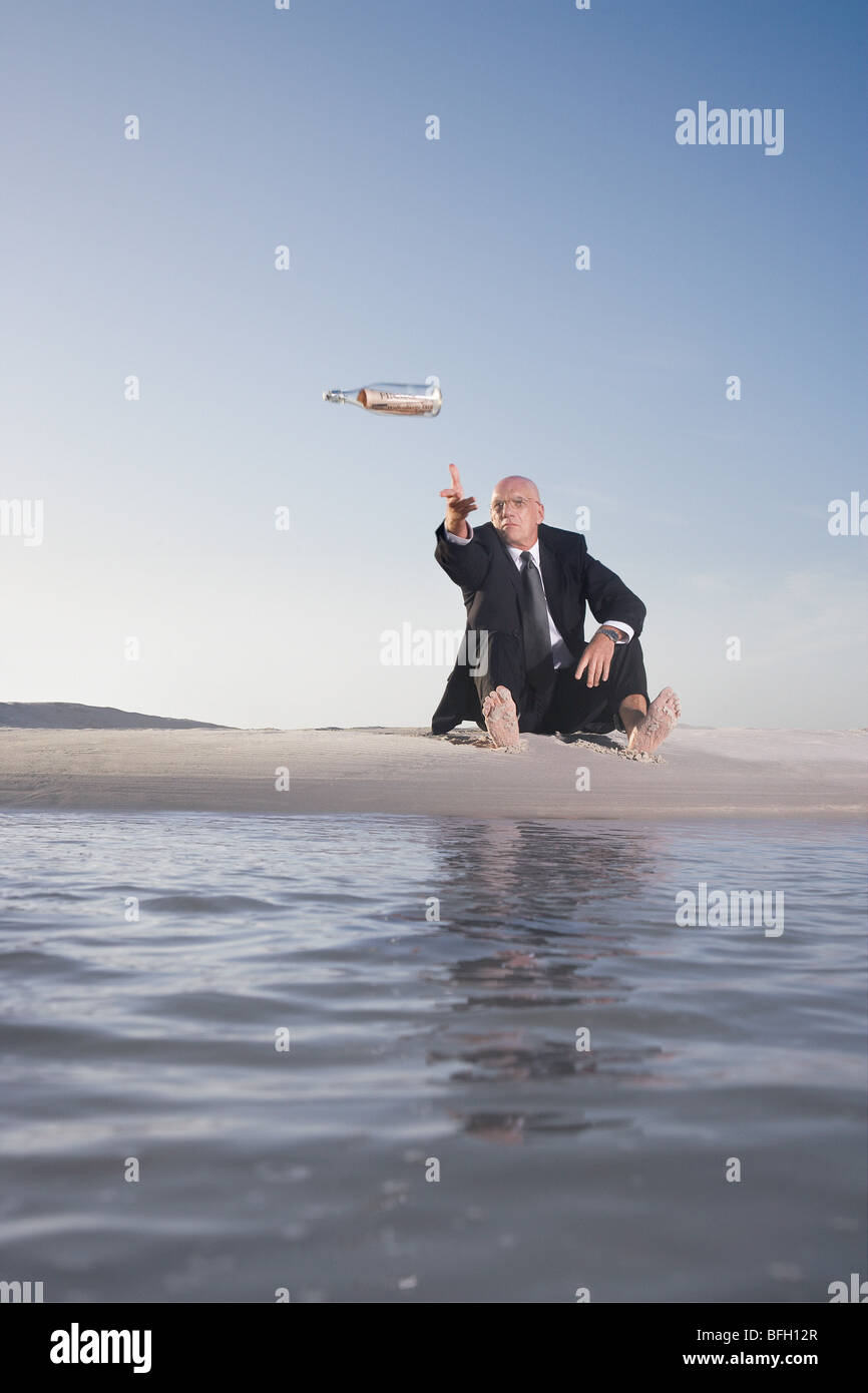 Business man sitting on beach, throwing message in bottle into sea, low