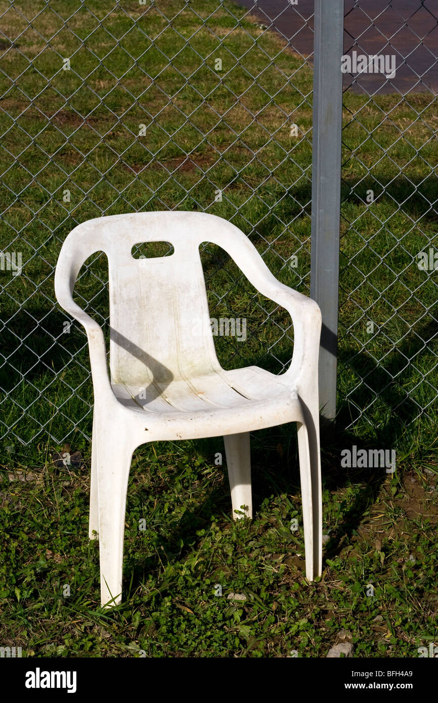 An empty dirty acrylic / plastic chair sitting in the grass in front of a chainlink fence. Stock Photo