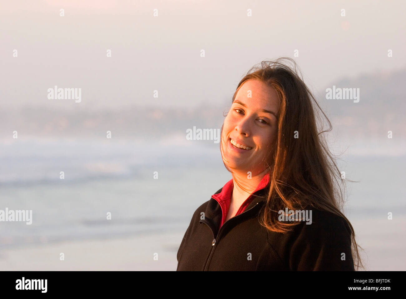 Head shot of a pretty young woman on a beach at sunset with her hair blowing in the wind and a big smile on her face. Stock Photo