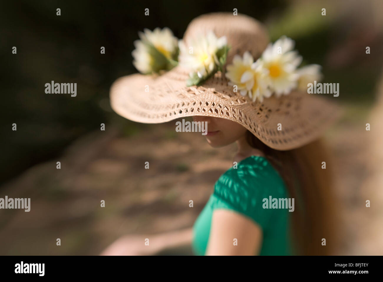 A long haired young woman or teen wearing a straw hat adorned with flowers sits in the sun on a summer day' Stock Photo