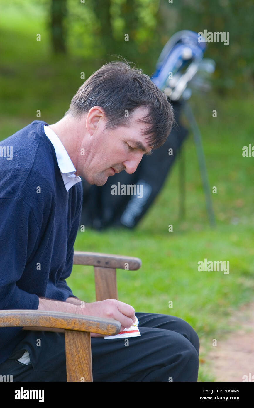 A middle aged white male playing golf filling out his score card after finishing a hole with his golf trolley in the background Stock Photo