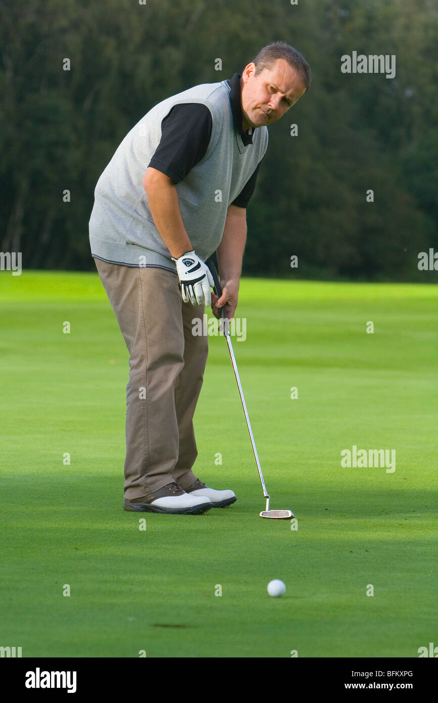 A middle aged white male playing golf watching his ball on its way to the hole on the green while holding his putter Stock Photo