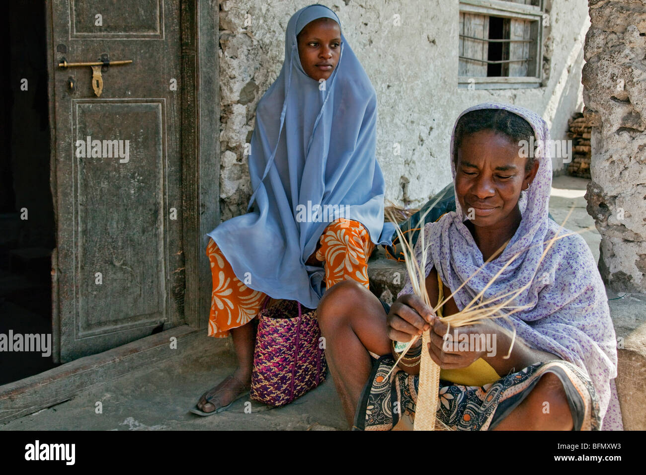 Tanzania, Zanzibar. Watched by her daughter, a Zanzibari woman weaves palm fronds to make baskets in Paje village. Stock Photo