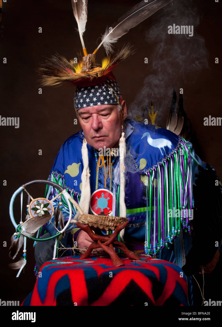 Native American Indian male in full regalia leaning over burning sage bowl with dream catcher in hand on brown background Stock Photo