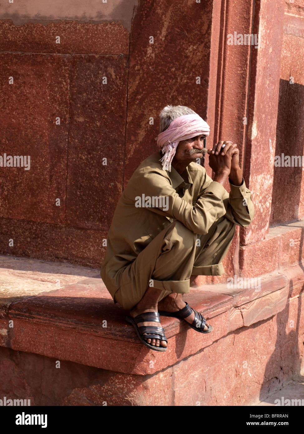 local man wearing turban and khaki clothes squats on the edge of a terrace at the Red Fort Agra with clasped hands Stock Photo