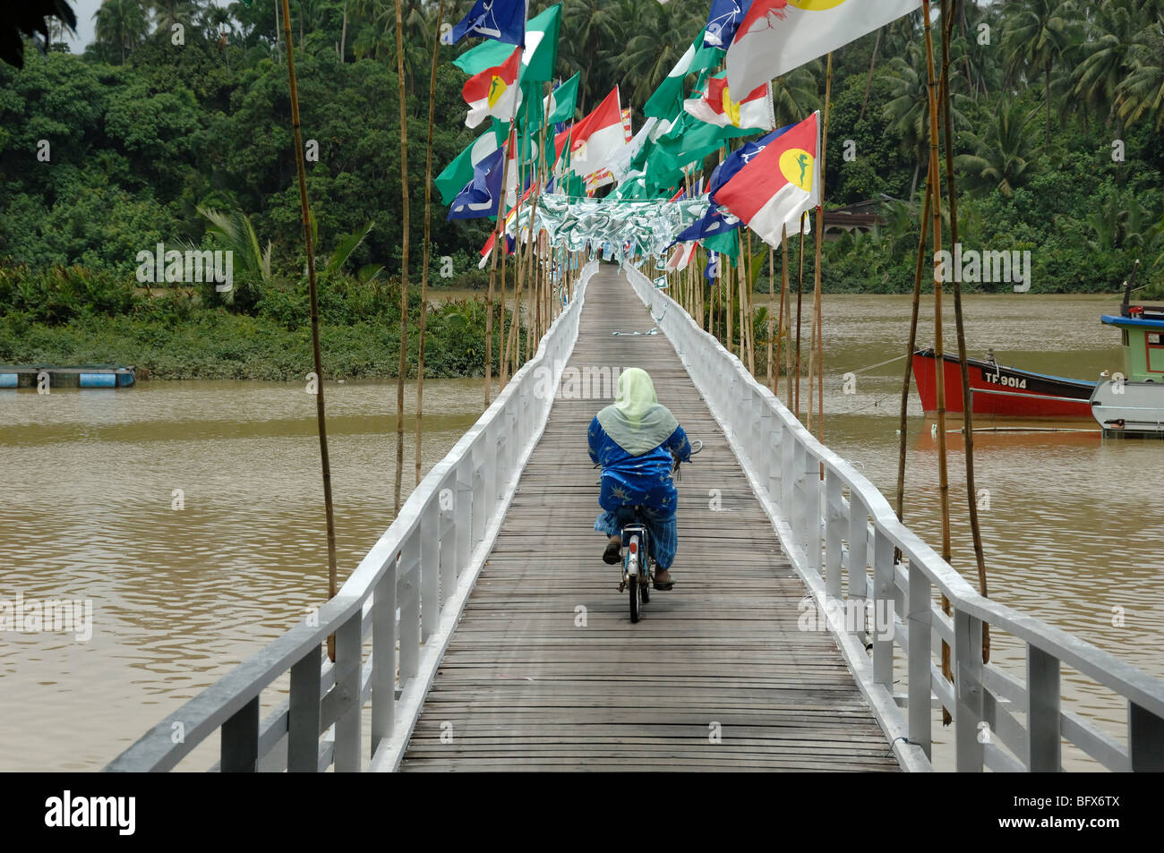 Malay or Malaysian Muslim Woman Cycling Across Wooden Foot Bridge Covered in Election Flags, Kuala Terengganu, Malaysia Stock Photo