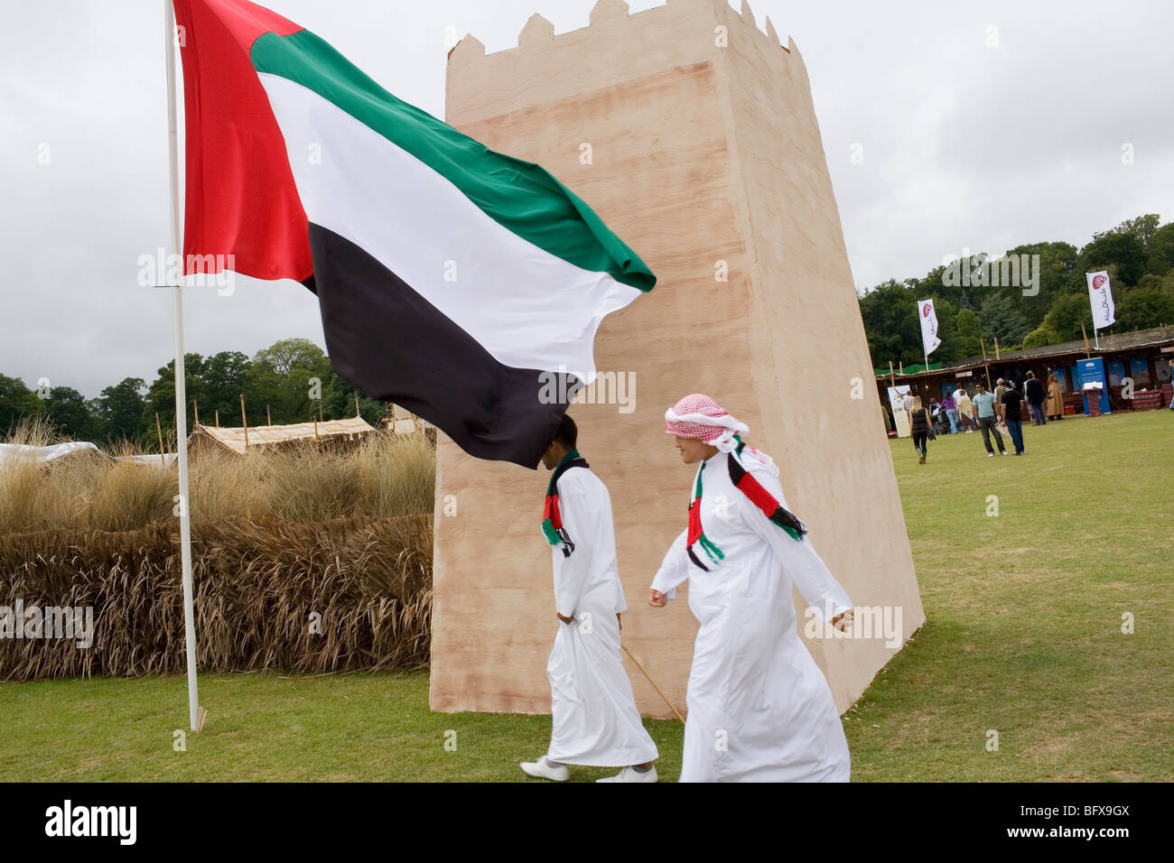 2 emirate men in traditional dress walk past their national flag flying at a falconr festival in the English countryside. Stock Photo