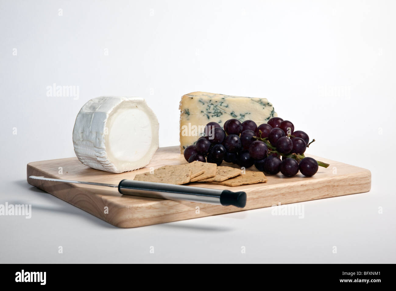 Stilton and goats cheese with red grapes and crackers on a wooden cheese board with cheese knife. Stock Photo