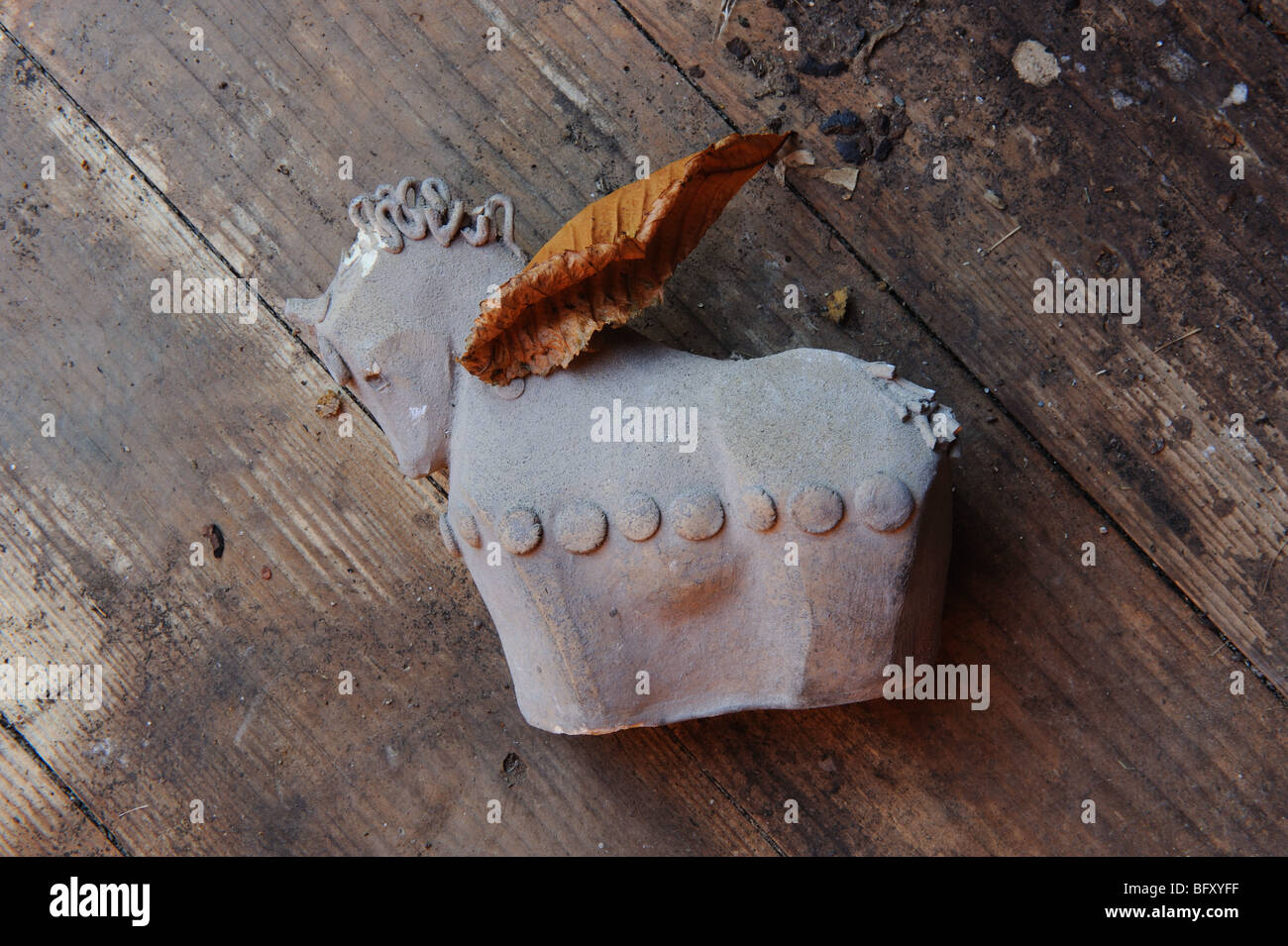 Unfinished, unglazed and broken ceramic horse ornament lies on dirty wooden floor. A golden leaf has fallen on it Stock Photo