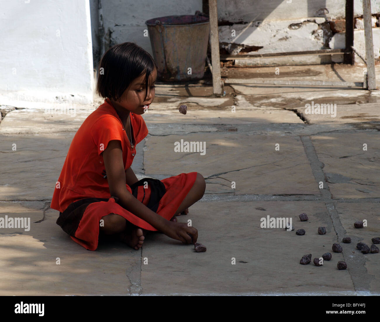 squatting girl dressed in red  playing jacks with stones on yard flagstones in India Stock Photo