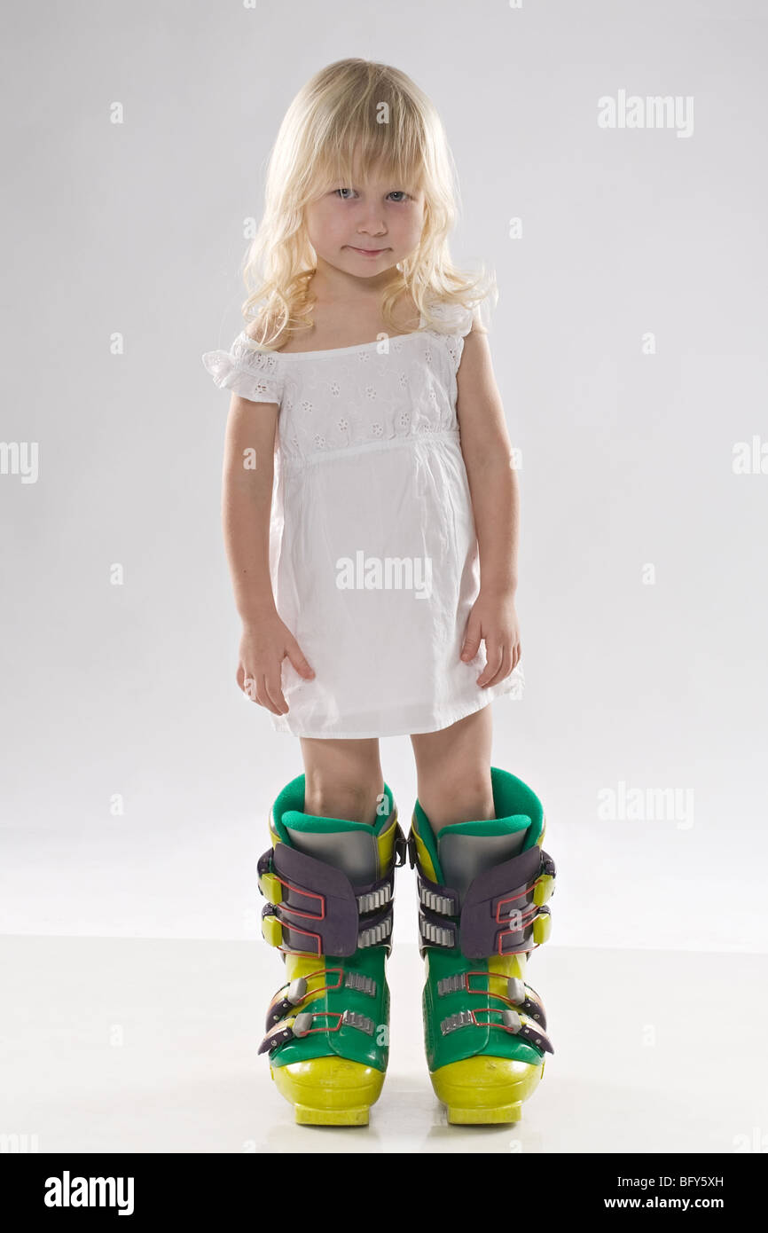 funny little girl in big mountain ski boots and short white dress stands on white background Stock Photo