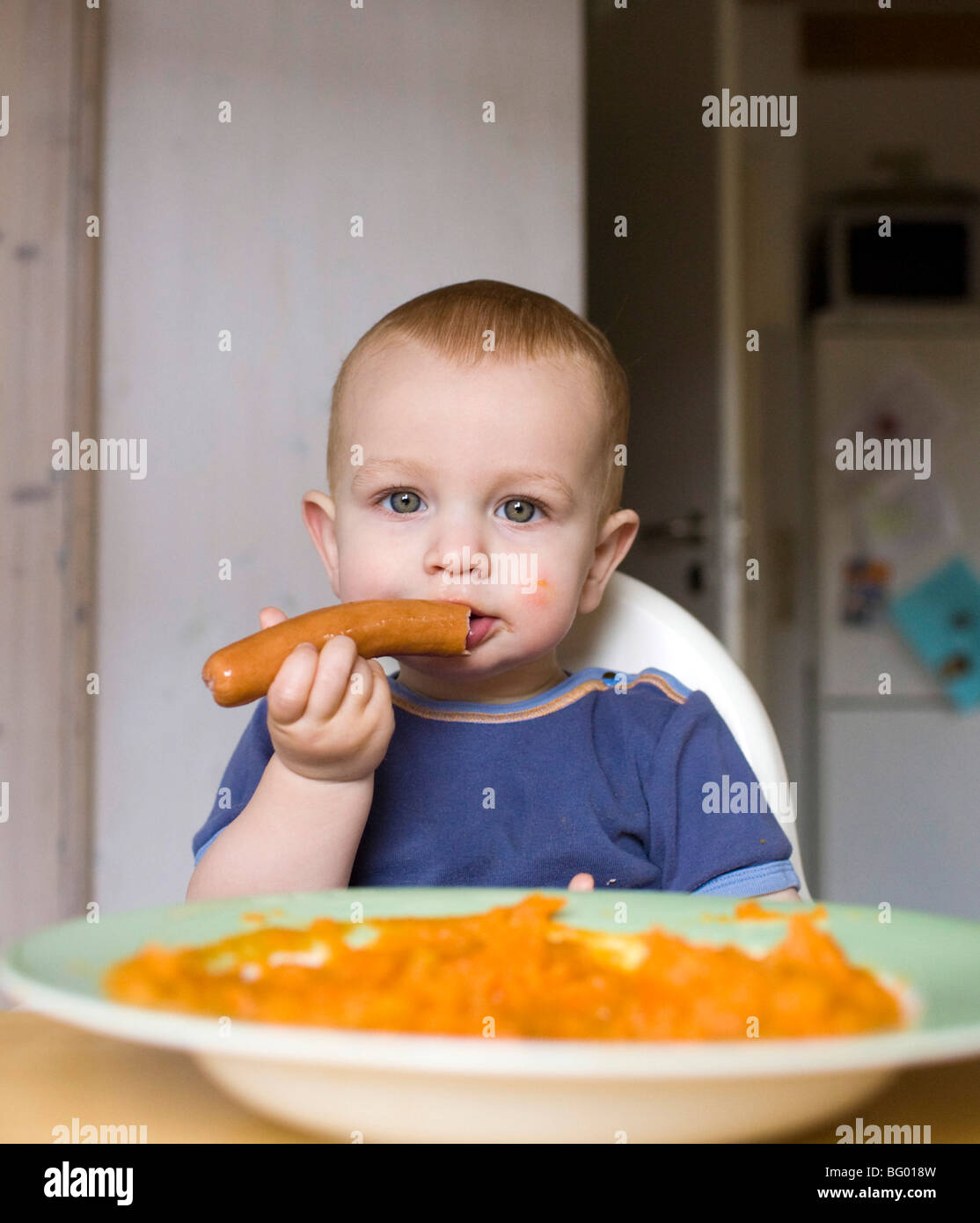 Young boy eating a sausage in a kitchen. Stock Photo