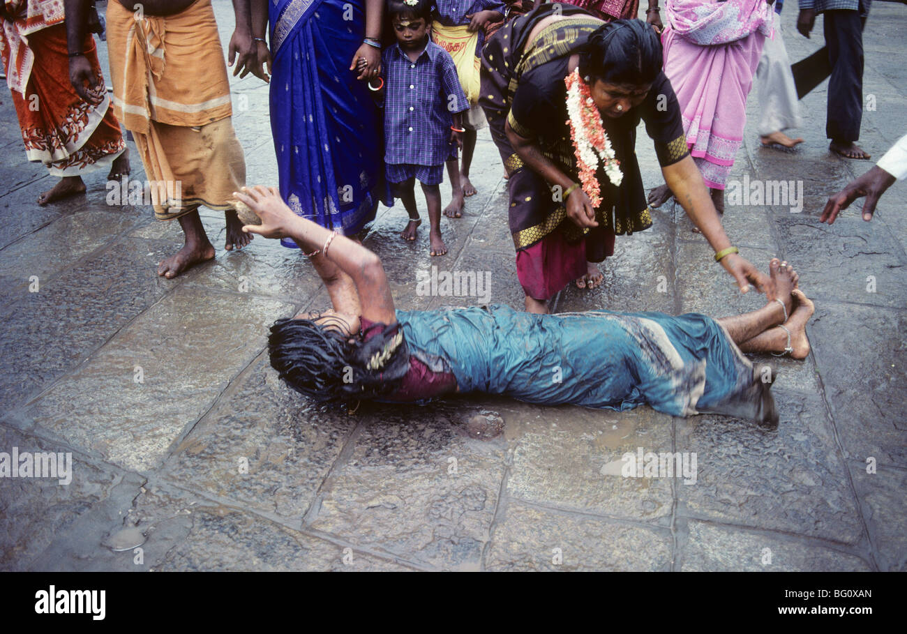 A pilgrim rolls on the ground which is hard pavement completely around the Palani Murugan Temple holding a coconut during the annual Hindu Thaipusam festival. She is assisted by family members who occasionally give her water. This is a form of kavadi which is an act of devotion represented by the hardship and pain she is experiencing. Stock Photo