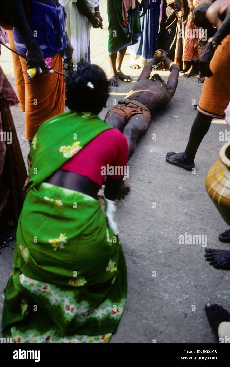 A pilgrim rolls on the ground which is hard pavement completely around the Palani Murugan Temple holding a coconut during the annual Hindu Thaipusam festival. He is assisted by family members who occasionally give her water. This is a form of kavadi which is an act of devotion represented by the hardship and pain she is experiencing. Stock Photo