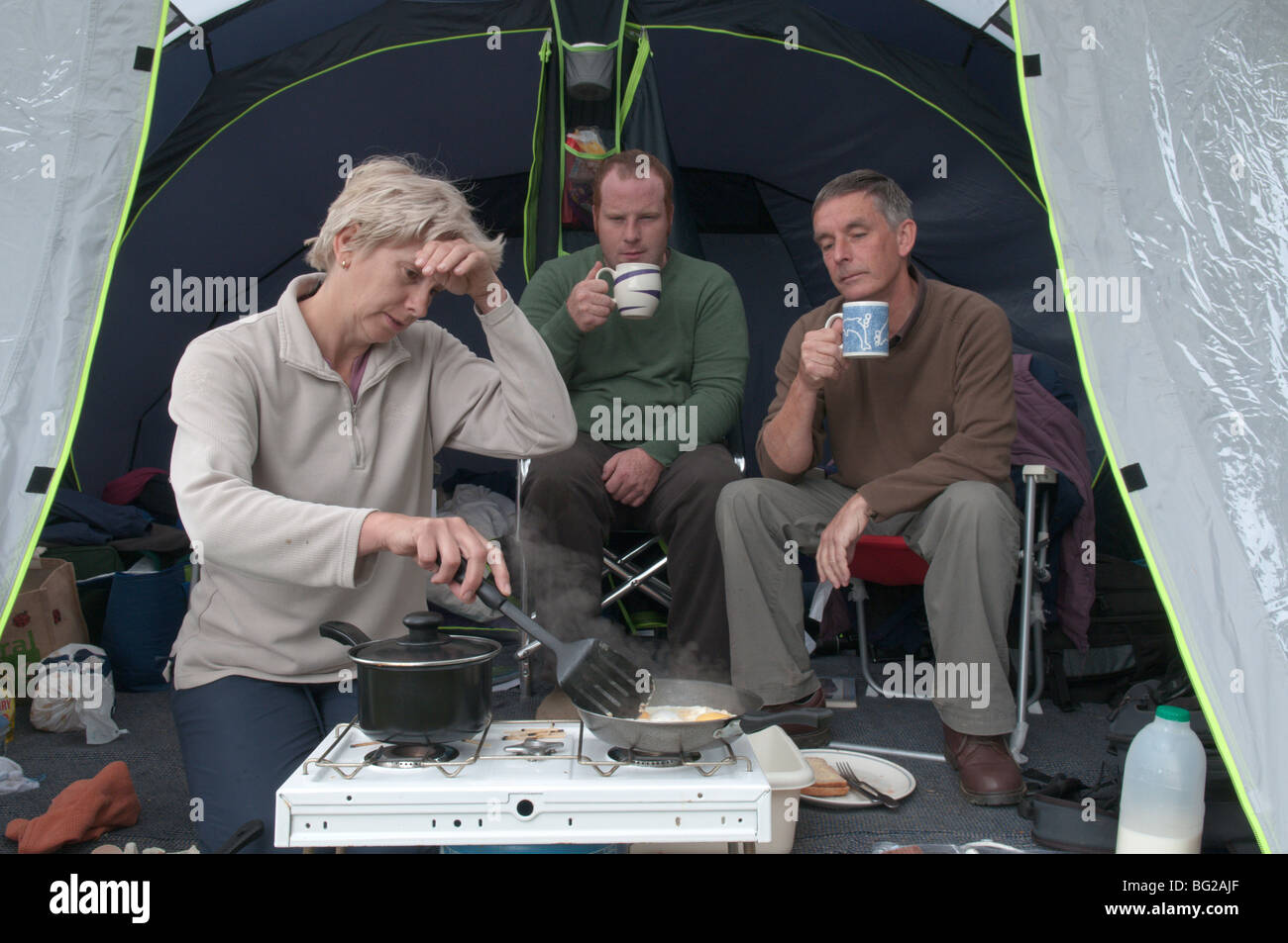 woman in tent cooking for two men as they just watch, Lazy men. Stock Photo