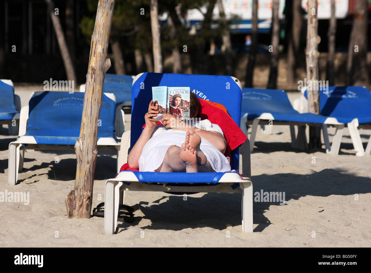 Lady relaxing in the sun on a beach lounger reading a book. Stock Photo