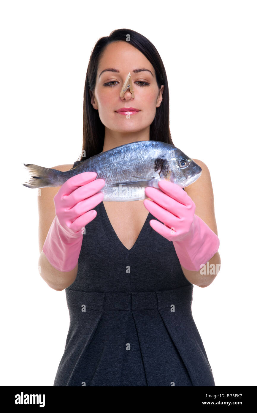 A woman wearing rubber gloves with a clothes peg on her nose holding a fish out in front of her, focus is on her face. Stock Photo