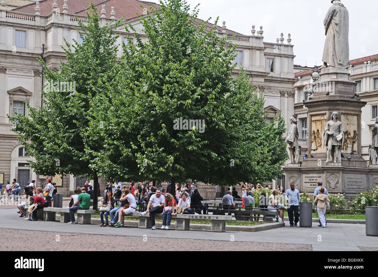 People relaxing on benches in Piazza dell Scala Milan Milano Italy Italia La Scala Opera House is behind the trees Stock Photo