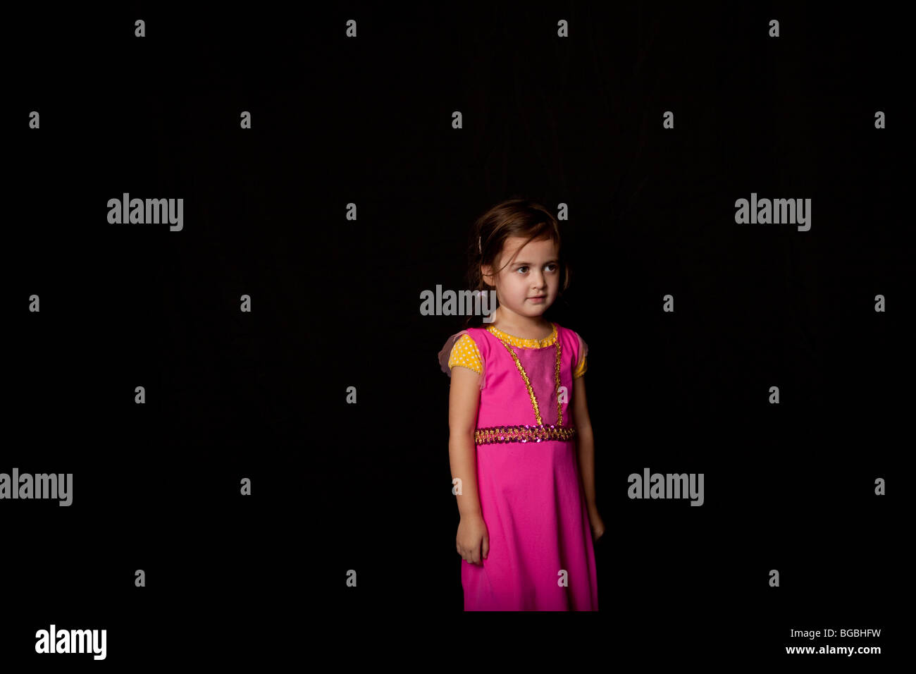 young girl standing alone against black background, wearing a pink dress. Stock Photo