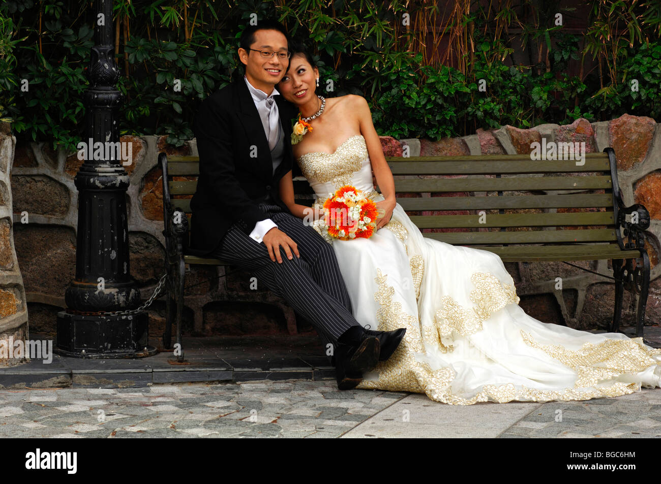 Chinese wedding couple in a long dress and a dark suit sitting on a bench, Hong Kong, China, Asia Stock Photo