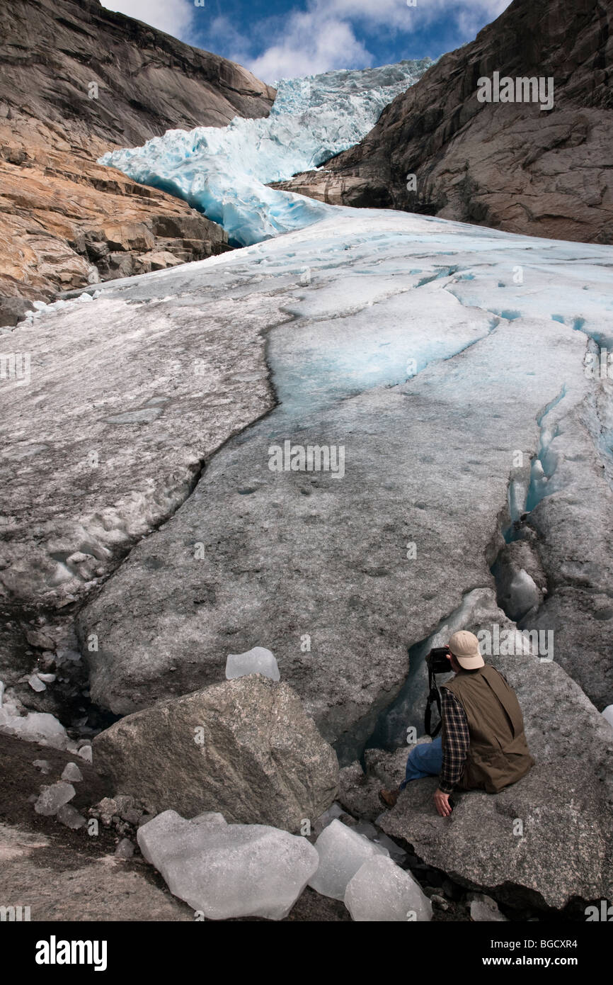 vertical landscapes with scale, male photographer sitting on melting ice base of Briksdal Glacier, Jostedal Park Norway in summer, Blue sky background Stock Photo