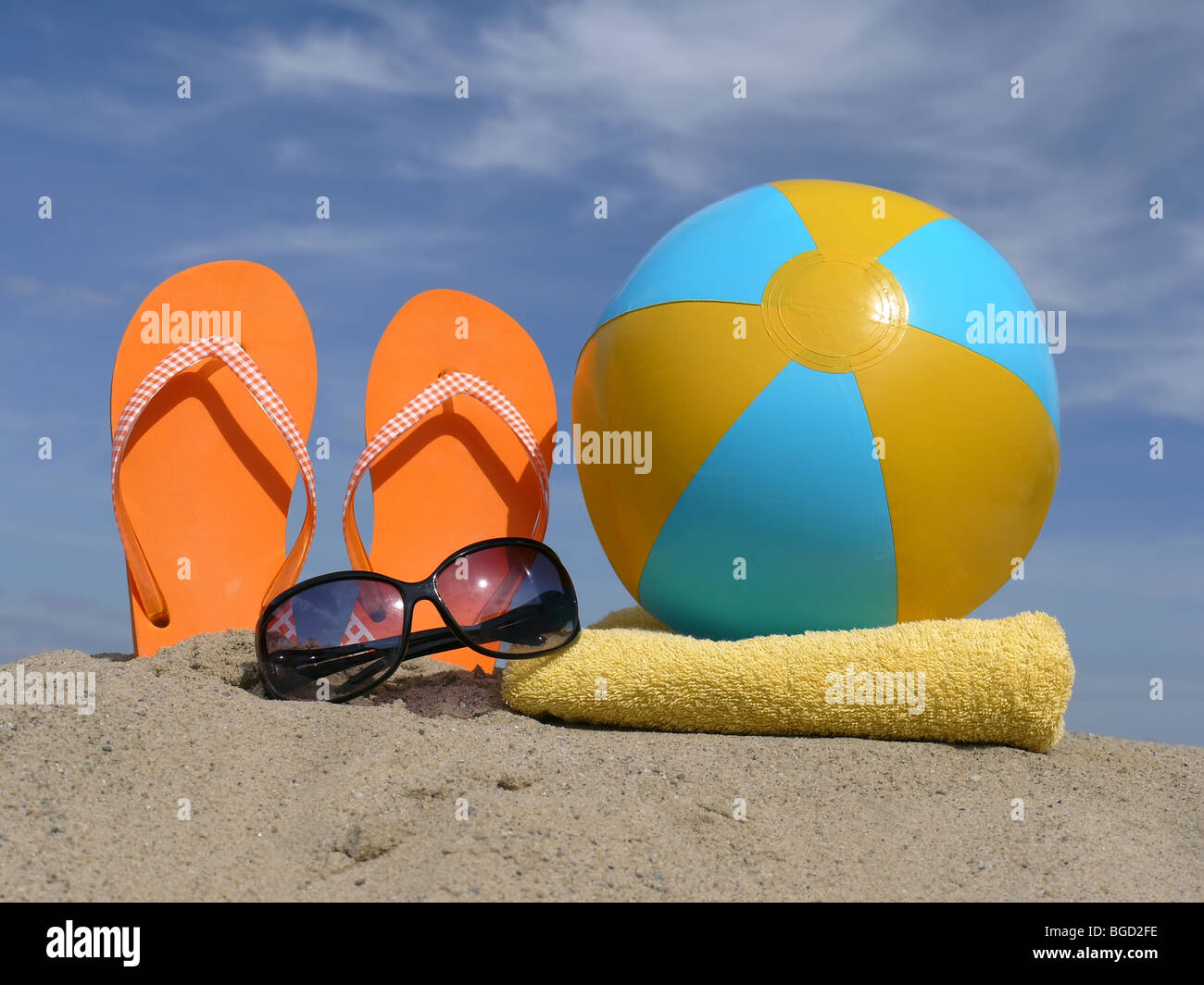 Orange flip-flops driven vertically into beach sand, sunglasses, inflated beach ball and  yellow bath towel over blue sky Stock Photo