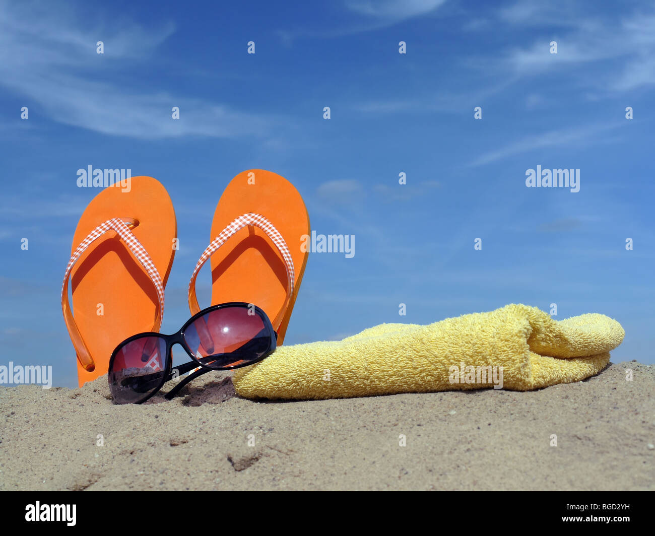 Orange flip-flops driven vertically into beach sand with sunglasses and yellow bath towel over blue sky Stock Photo