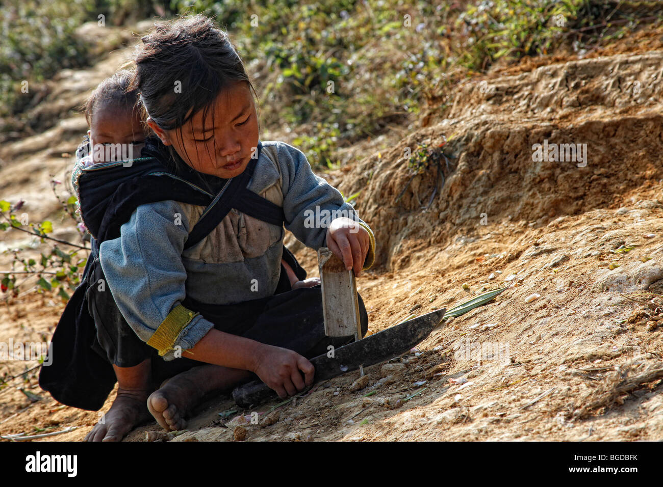 Small child with a baby on her back, sitting on a trail in the mountains of North Vietnam near the mountain town of Sa Pa, Viet Stock Photo