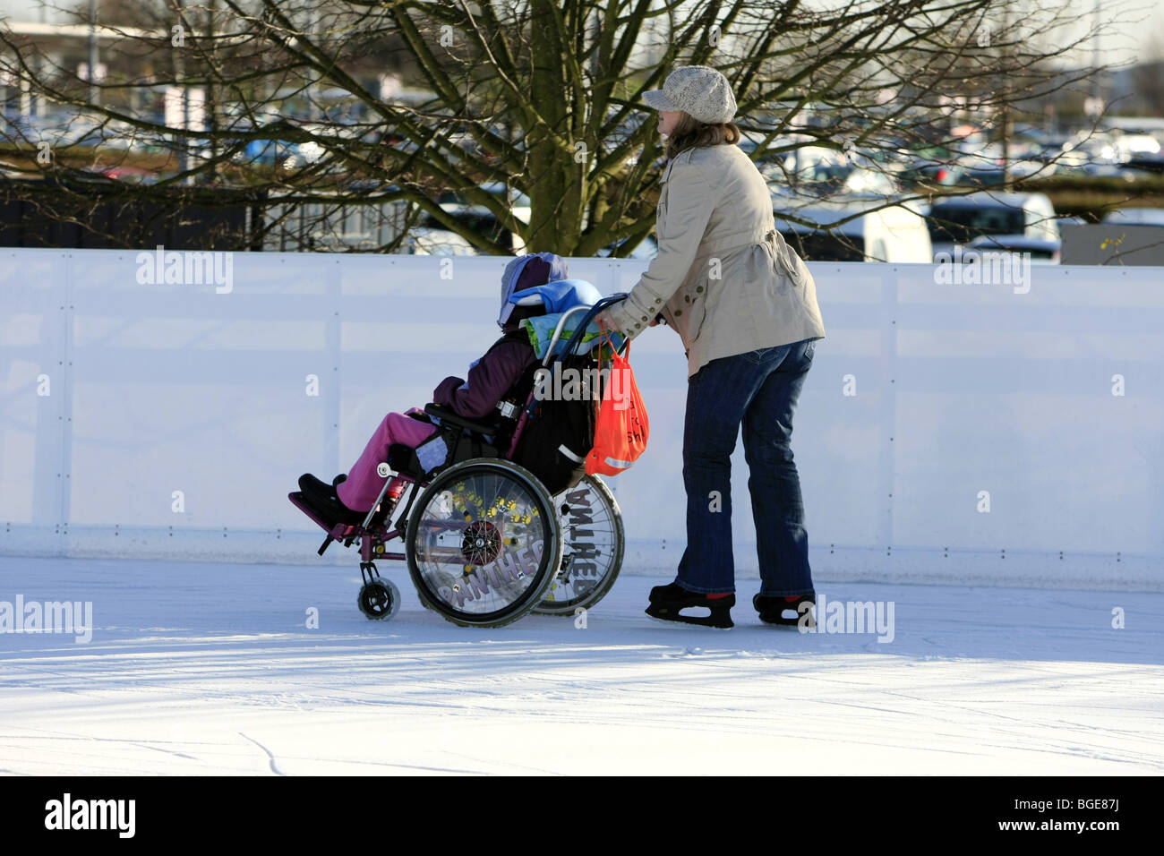 Disabled child in a wheelchair being pushed around an ice rink by a carer on a winter day out Stock Photo
