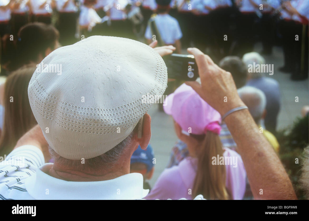Rear view from above of elderly man wearing flat white cap taking photo on compact digital camera of youth brass band Stock Photo