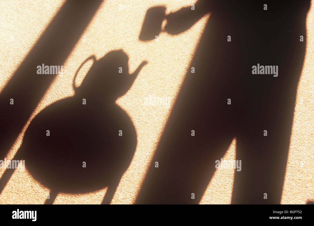 Shadows on warm background of figure holding mug and standing by small circular table with teapot on it Stock Photo