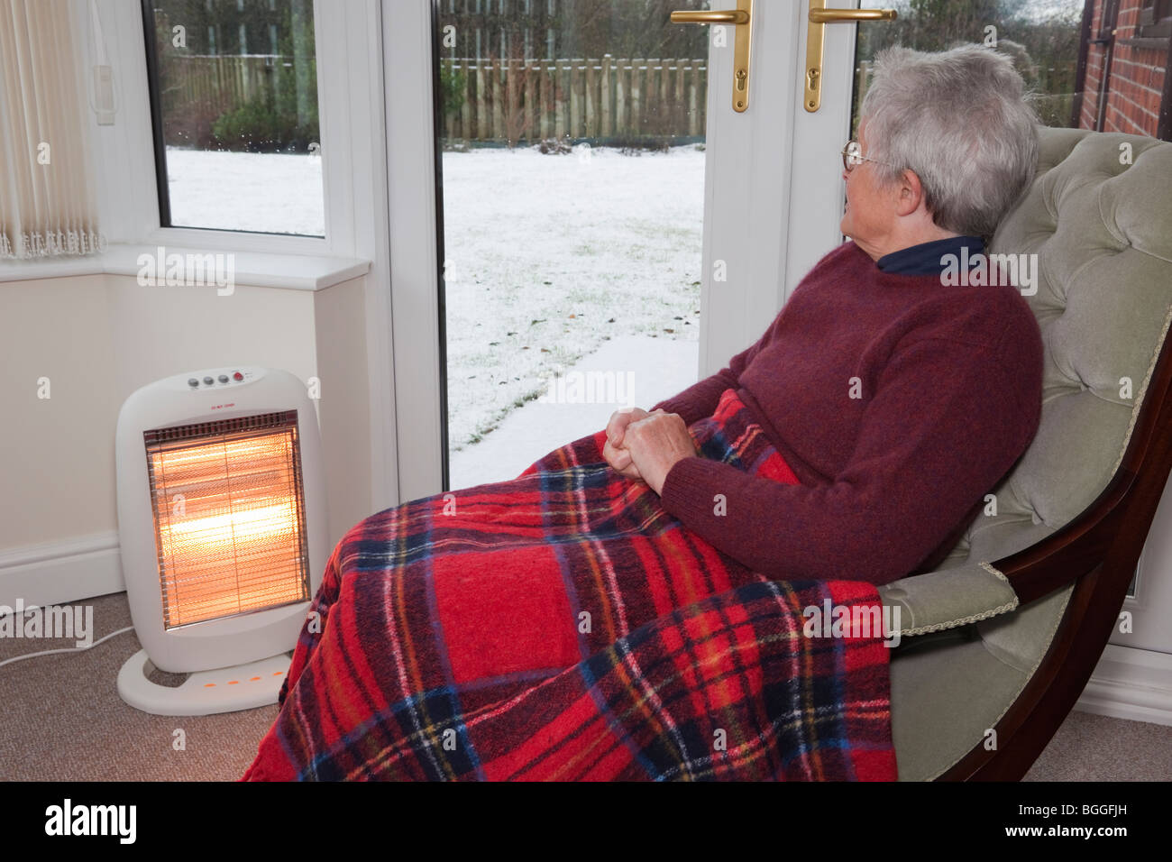 Elderly old woman alone at home with a rug on knees keeping warm indoors by electric fire with snow outside the windows in winter UK Britain Stock Photo