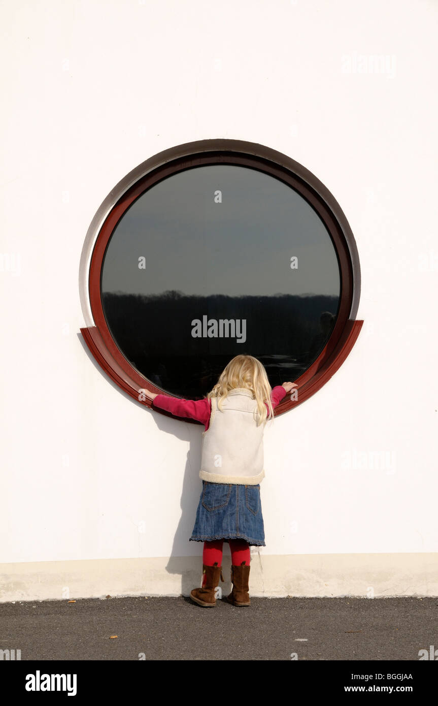 Stock photo of a 3 year old girl peeking into a round window. Stock Photo