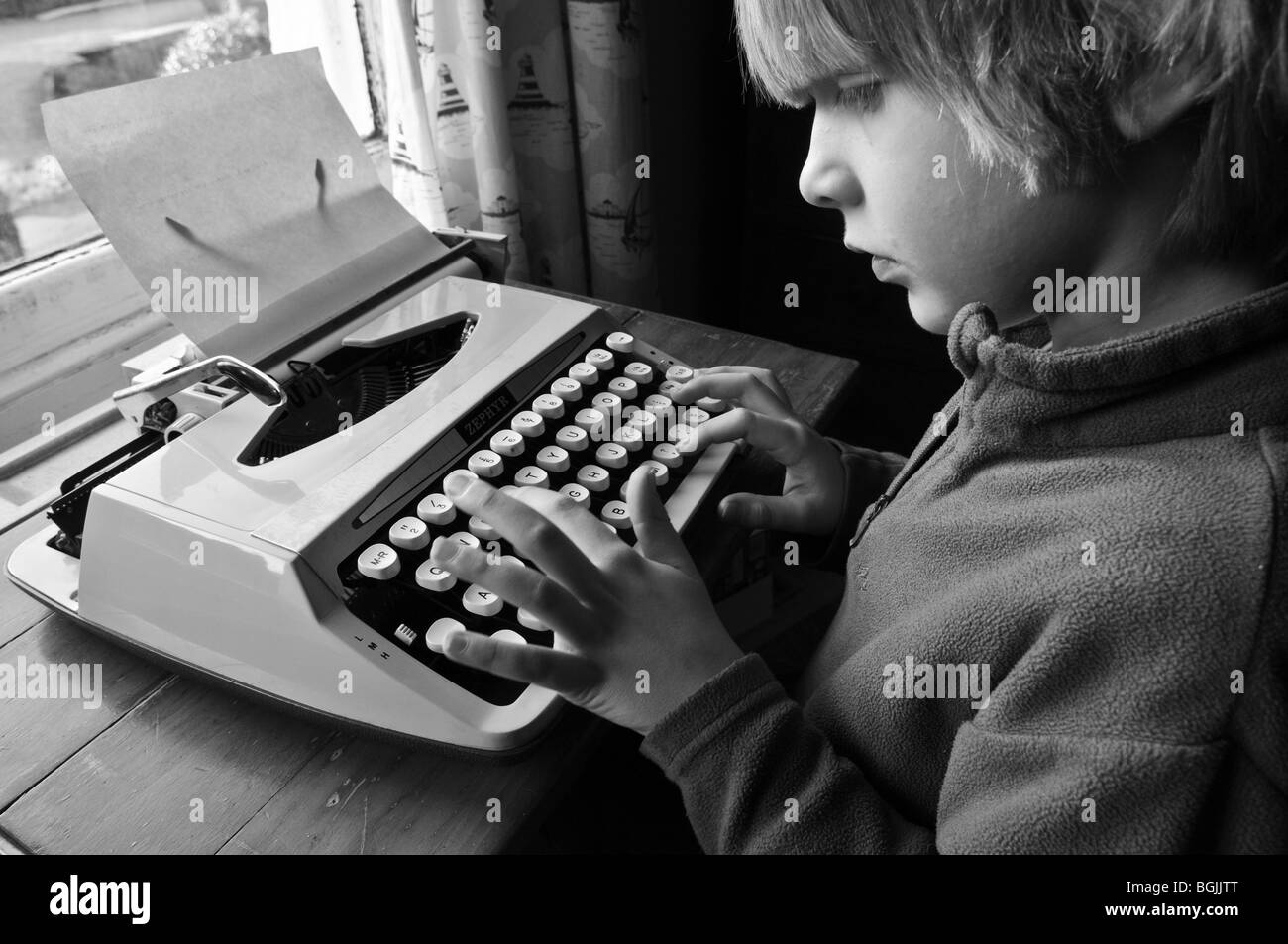 Young boy sitting at desk by the window using a typewriter, pressing keys to type a letter. Stock Photo
