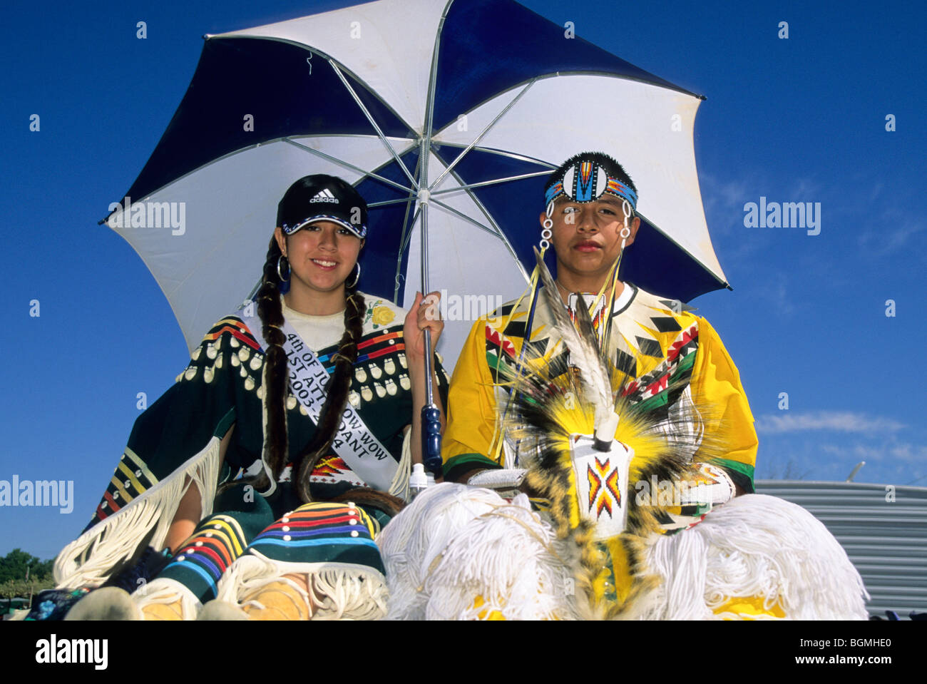 Two teenagers dressed in traditional regalia under an umbrella during a