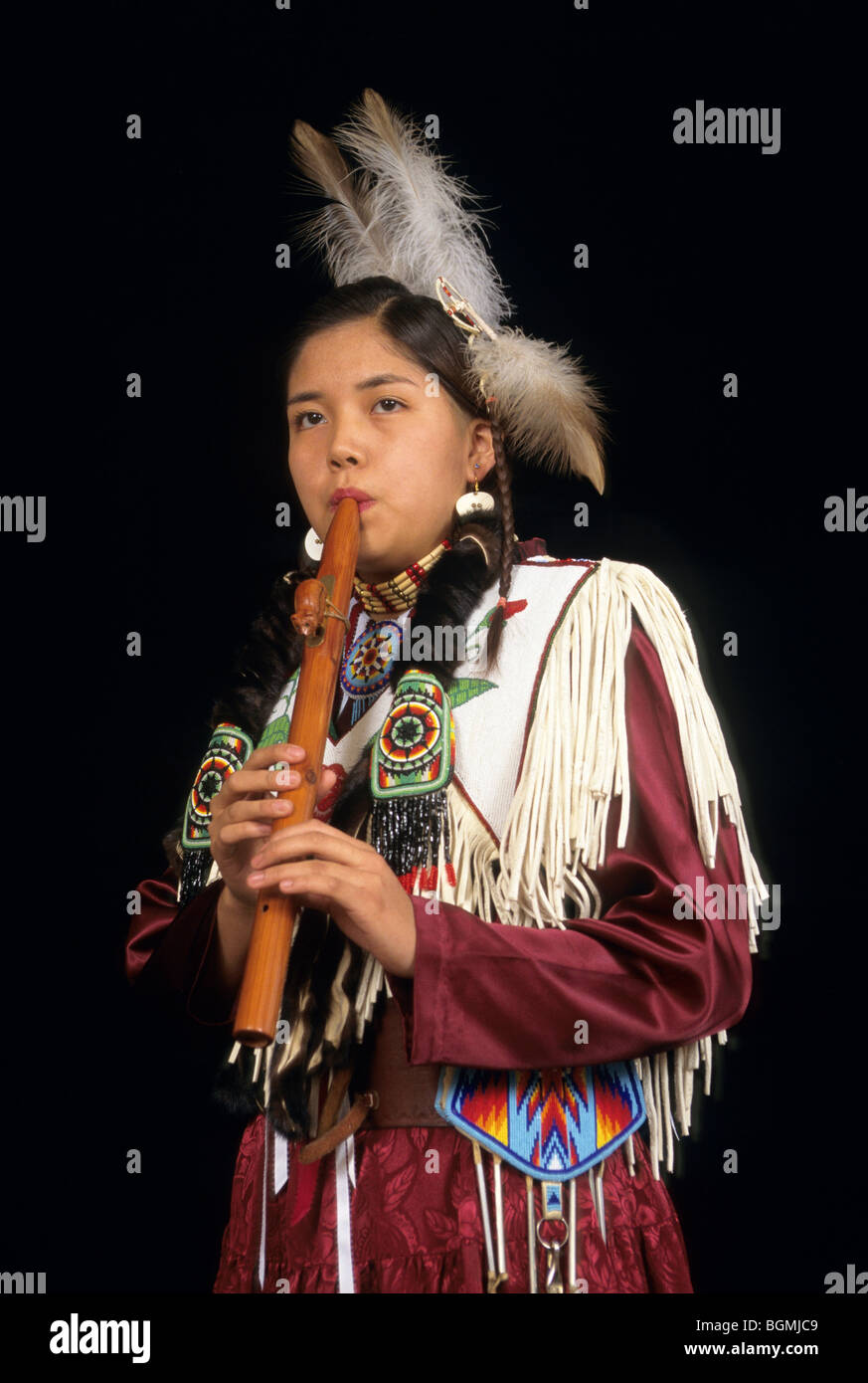 Native American teenage girl dressed in traditional dress decorated with beadwork plays traditional wooden flute Stock Photo