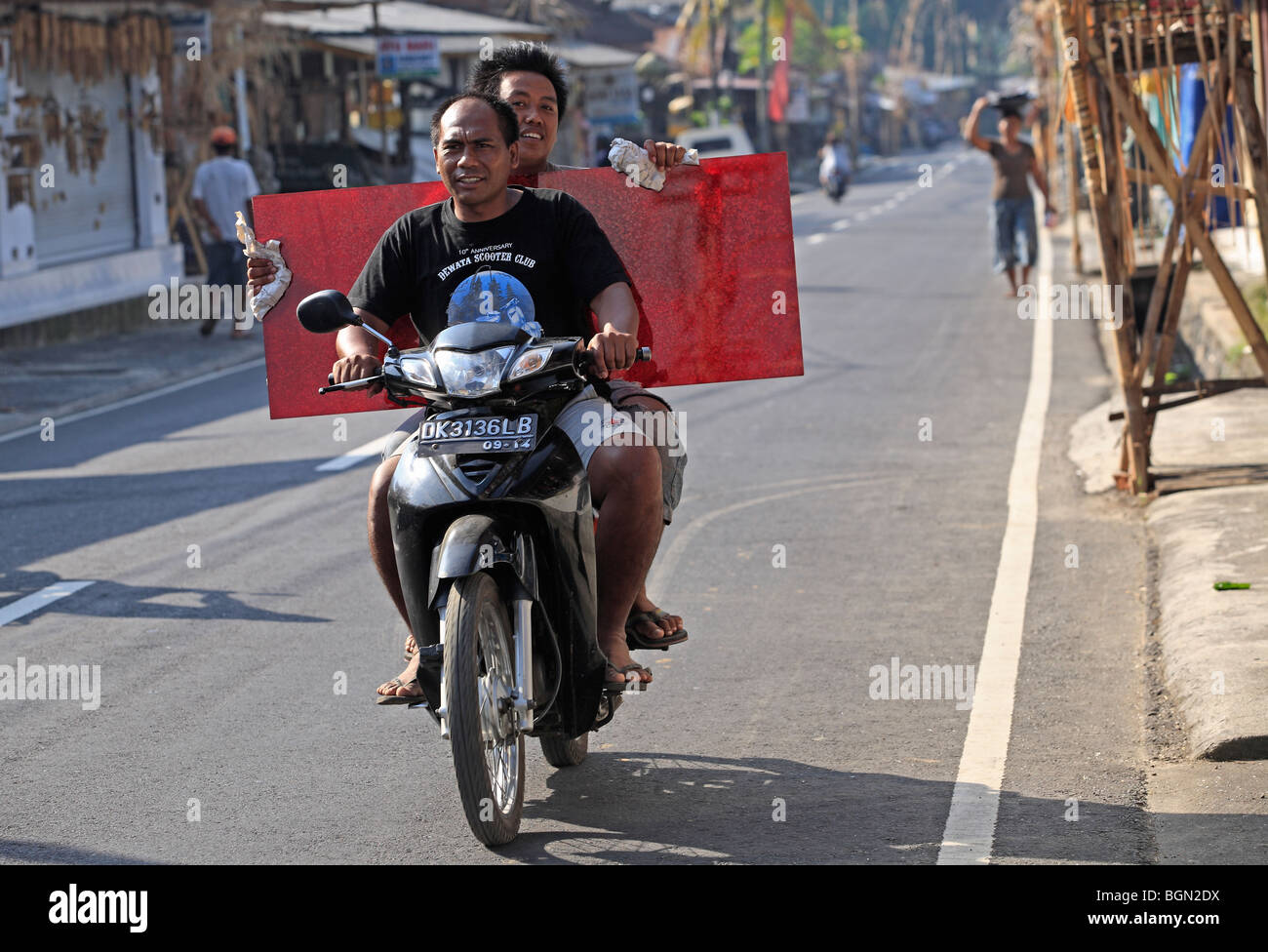 Two men riding on a motorcycle, one carrying a large red item.Tegallelang Village, Ubud, Bali, Indonesia Stock Photo