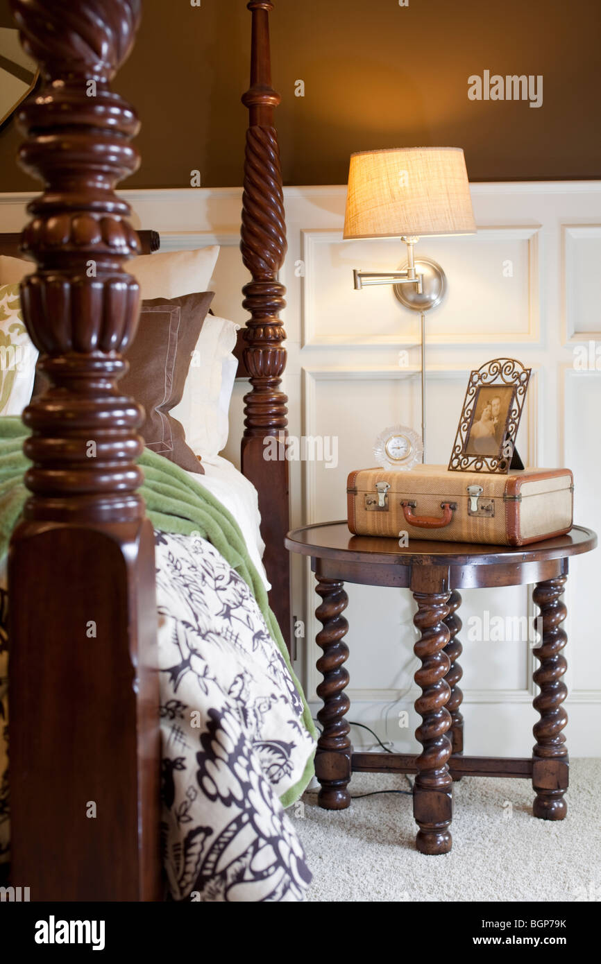 Master bedroom with carved wooden four poster bed, bedside table with suitcase and lamp. Stock Photo