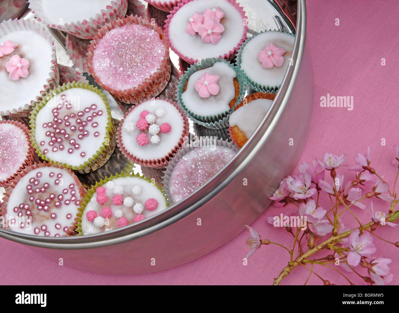 Silver cake tin with decorated fairy cakes on pink background with cherry blossom Stock Photo