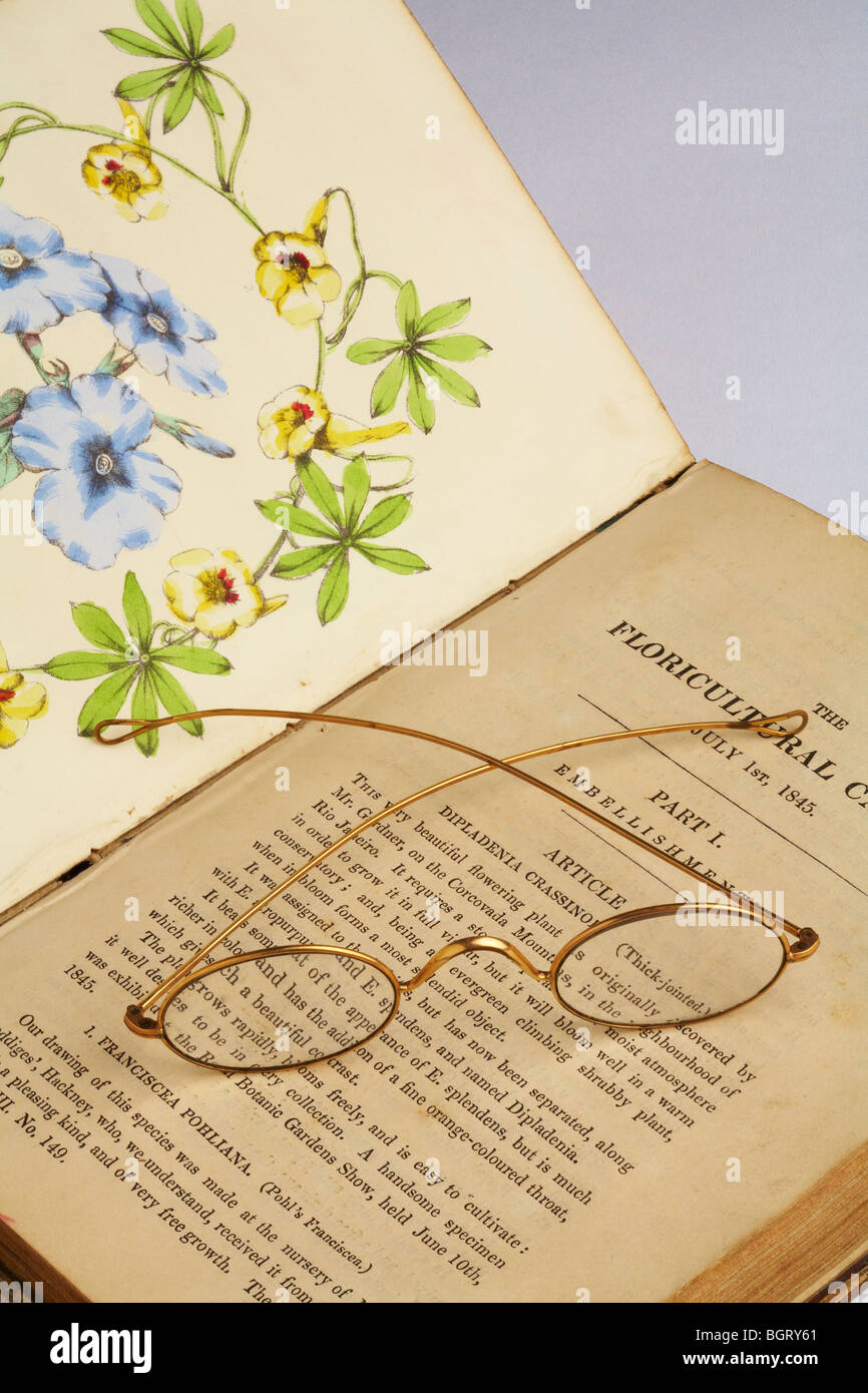 Pair of antique gold rimmed spectacles laying across open antique book on horticulture hand painted colour picture Stock Photo