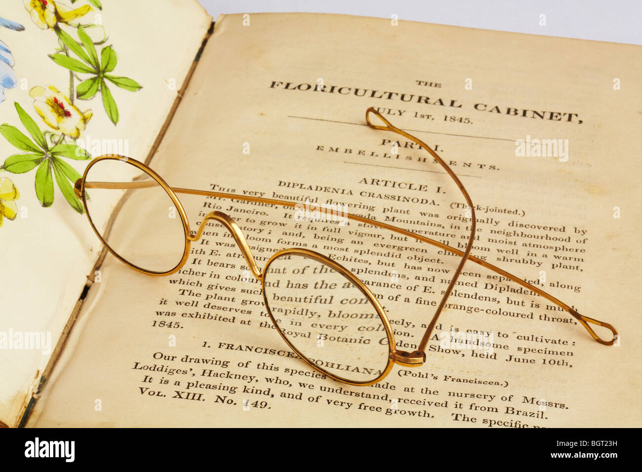 Pair of antique gold rimmed spectacles laying across open antique book on horticulture hand painted colour picture Stock Photo