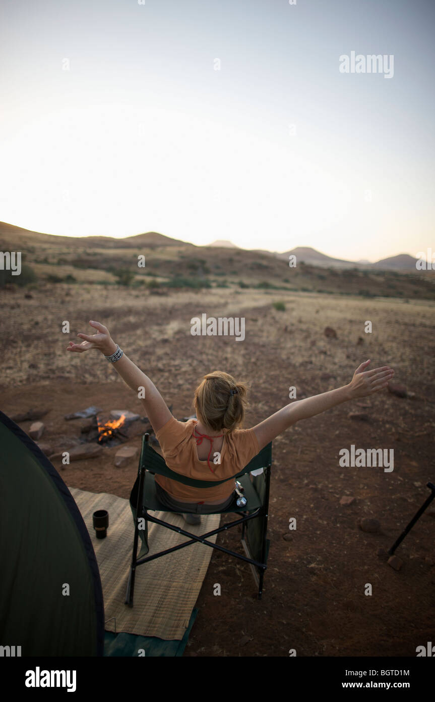 Rear view of woman at sunset at camp fire with arms in the air, Sesfontein, Kaokoland, Namibia Stock Photo