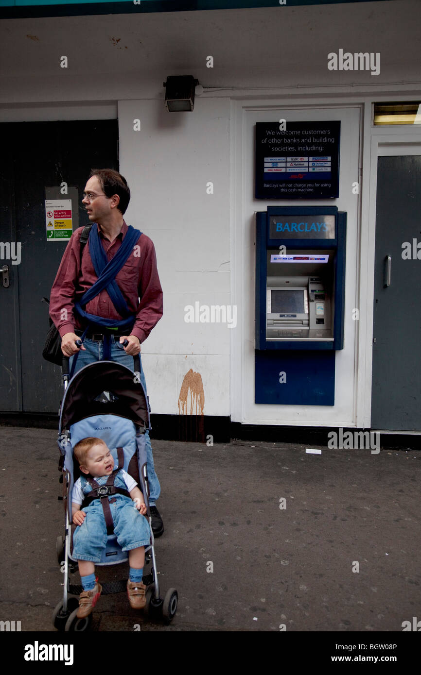 Father with baby pushes his child in a pram away from a cashpoint ATM machine. Stock Photo